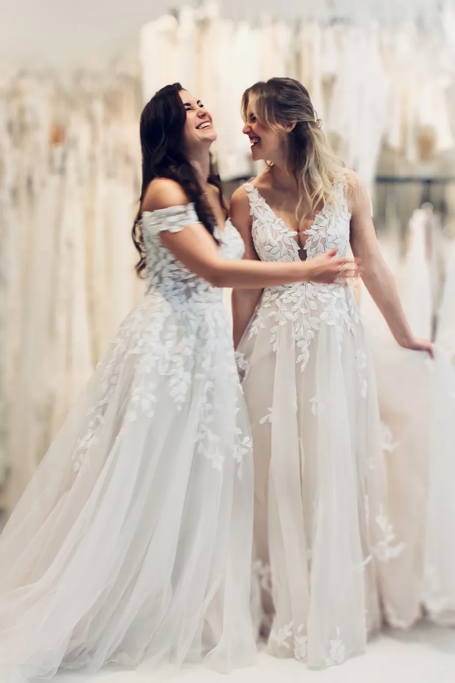 zwei ähnliche Brautkleider mit Blätterspitze an zwei unterschiedlichen Frauen, die sich anlachen und viel Spaß beim Fotoshooting haben